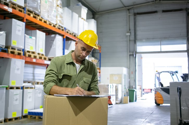 serious-male-logistic-engineer-hardhat-overall-standing-stack-boxes-completing-documents-copy-space-blue-collar-labor-concept